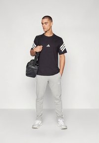 Černé tričko s bílým logem, šedé tepláky s bílými pruhy a bílé tenisky. Modelka nese černou kabelku na rameno.