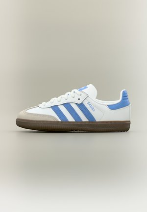 Fehér Adidas Samba edzőcipő kék csíkokkal és sarokkal, bézs orrborítással és barna gumitalppal, semleges háttéren.