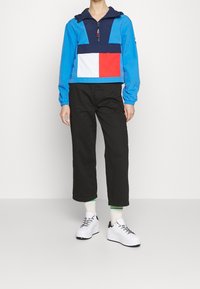Cropped blue windbreaker with navy hood and multicolored chest panel (white, red). Paired with black wide-leg pants and white sneakers.