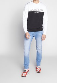Felpa a blocchi di colore in bianco e nero con testo "Calvin Klein", abbinata a jeans azzurri chiari e sneakers a fantasia rossa.