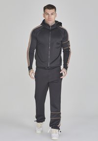 Chándal atlético negro con sudadera con cremallera y pantalones ajustados. Presenta franjas laterales doradas y naranjas. El material parece suave y ajustado.