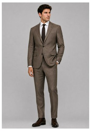 Homme portant un costume marron ajusté avec une chemise blanche, une cravate sombre et des chaussures habillées marron foncé, debout avec une main dans la poche.