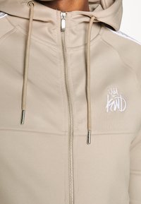 Beige zip-up hoodie med dragsko i huvan, silverfärgad dragkedja och broderad logotyp. Slät textur med sidopanelaccent.