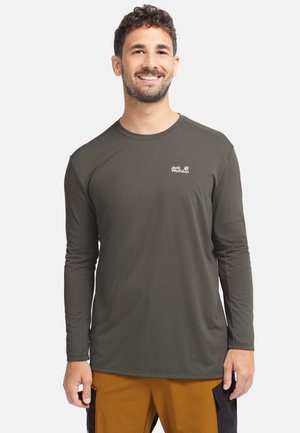 Lachende man met een olijfgroen Jack Wolfskin shirt met lange mouwen en mosterdgele broek met zwarte zijzakken, staand tegen een witte achtergrond.