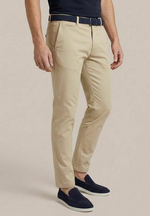 Man die beige slim-fit broek, marineblauwe riem en marineblauwe slip-on schoenen draagt, staand tegen een neutrale achtergrond.