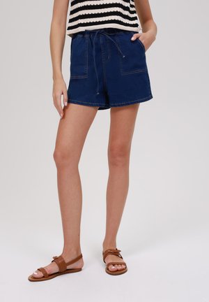Shorts en denim bleu foncé avec une taille élastique et un cordon de serrage, comportant deux poches avant et un ourlet replié. Sandales à lanières marron.