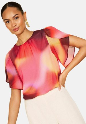 Vrouw die een kleurrijke doorschijnende blouse met vloeiende korte mouwen en lichte broek draagt, poseert met één hand op haar heup.