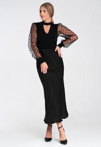 Schwarzes Kleid mit taillierter Blende und transparenten Punkteärmeln, V-Ausschnitt und einem fließenden Rock. Getragen mit schwarzen High Heels.