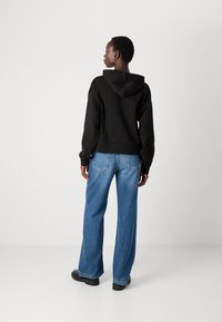 Sweatshirt noir avec capuche, offrant une coupe décontractée. Associé à un jean bleu à jambes larges et des chaussures à plateforme noires. Design minimaliste.