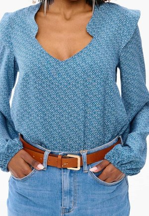 Nainen, jolla on sininen kuviollinen pitkähihainen paita, joka on tungettu korkeavyötäröisten farkkujen sisään ja ruskea vyö, kädet farkkujen taskuissa lepäämässä.