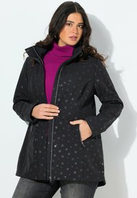 Žena oblečená v černé bundě se vzorem hvězd přes fialový rolák, s jednou rukou v kapse bundy a druhou si upravující zip.