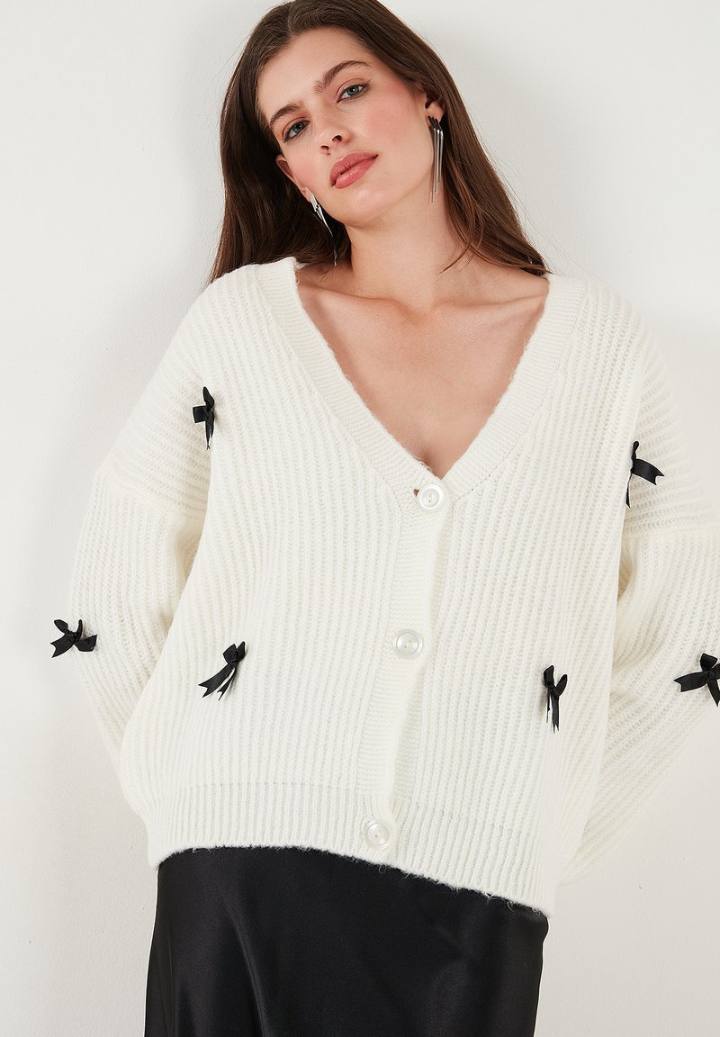 Kermanspun ribbinnitteinen cardigan, jossa on V-kaula-aukko, nappikiinnitys ja mustat nauhaaksentit hihoissa. Pehmeä rakenne ja rento istuvuus.