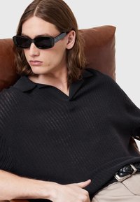 Svart stickad polo med V-ringning och korta ärmar, med ribbad textur. Modellen bär rektangulära svarta solglasögon och sitter på en brun lädersoffa.