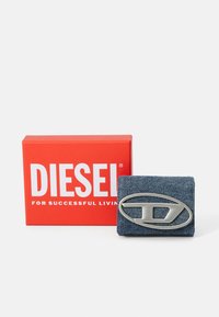 Džínová peňaženka so striebornou logom doskou, prezentovaná v červenej krabičke s nápismi "DIESEL" a "PRE ÚSPEŠNÝ ŽIVOT" potlačenými v bielej farbe.