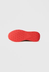 Czerwona gumowa podeszwa buta z teksturowanym wzorem diamentu i wytłoczonym logo na środku, z gładką obwódką.