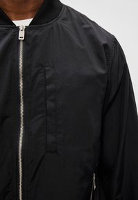 Schwarze Bomberjacke mit glatter Textur, geripptem Kragen, Frontreißverschluss und einer Seitentasche. Minimalistisches Design mit eleganten Akzenten.