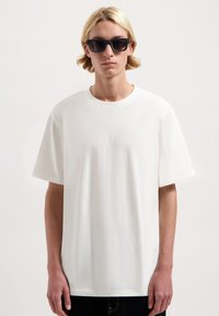 Weißes Baumwoll-T-Shirt mit lockerer Passform, kurzen Ärmeln und dezent eingeprägtem Logo auf der Brust. Getragen mit schwarzen quadratischen Sonnenbrillen.