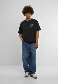 Svart bomull T-skjorte med et blått grafisk design på brystet. Kombinert med løstsittende blå jeans og nøytrale joggesko.