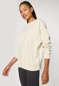 Vaalean beige collegepaita, jossa on pyöreä kaulus ja ribbireunat, pieni violetti logo rinnassa. Tasainen rakenne, rento istuvuus.