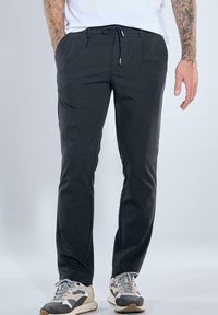 Man wearing black casual pants with drawstring waist and white, black, and beige sneakers, standing against a plain light background.