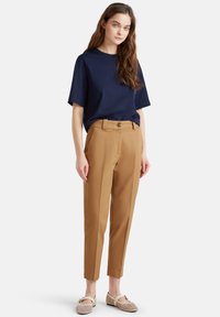 Młoda kobieta z długimi brązowymi włosami, ubrana w granatową koszulkę, beżowe spodnie z wysokim stanem i kremowe płaskie buty z wycięciami.