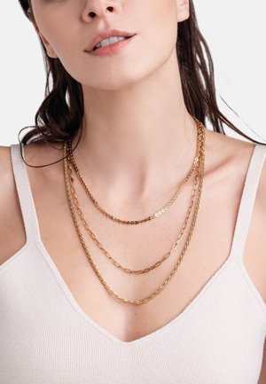 Collane in oro con strati di diverse stili di catene—anelli, texturizzato e liscio—indossate sopra una canotta a coste di colore chiaro.