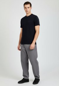 Maglietta nera a maniche corte con scollo rotondo, pannelli laterali strutturati, abbinata a pantaloni grigi e sneakers nere su uno sfondo neutro.