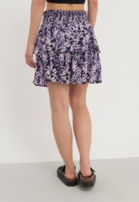Jupe noire à volants avec un motif floral violet et blanc, taille élastique, avec un design en couches. Associée à des chaussures compensées noires.