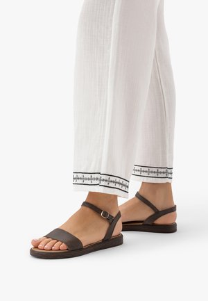 Stopy w ciemnobrązowych płaskich sandałach z paskami wokół kostki, w połączeniu z białymi spodniami z czarnym haftowanym wykończeniem.