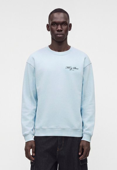 Homme portant un sweat-shirt bleu clair avec le texte "Filling Pieces" sur la poitrine gauche et un pantalon sombre sur un fond blanc uni.