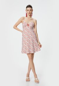 Witte zomerjurk met rode bloemenprint en groene bladeren, voorzien van een V-hals, spaghettibandjes en een uitlopende rok. Rode kwastoorbellen maken de outfit af.