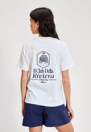 Kobieta o długich ciemnych włosach, ubrana w białą koszulkę z nadrukiem i granatowe szorty, stoi tyłem na tle jednolitego jasnego tła.