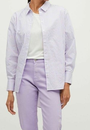 Kobieta ubrana w biały T-shirt z okrągłym dekoltem, koszulę zapinaną na guziki w pionowe paski w kolorach lawendowym i białym oraz jasnofioletowe spodnie z wysokim stanem.