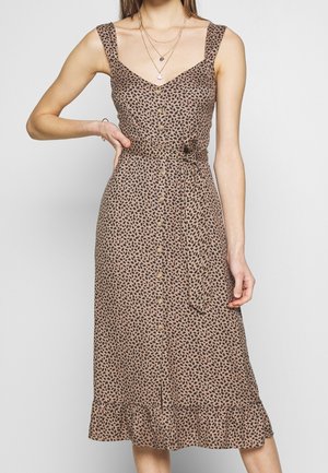 Robe midi marron sans manches avec motif feuilles noires, boutons au devant, ceinture à la taille et ourlet à volants portée par une personne avec des colliers superposés et un bracelet.