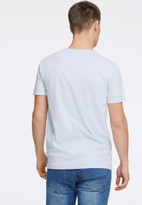 Vaaleansininen puuvillapaita, jossa on pyöreä kaula-aukko ja lyhyet hihat, nähtynä takaa. Yhdistetty sinisiin farkkuihin.