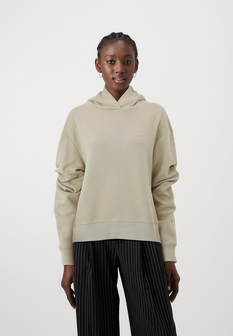 Felpa beige chiara con una vestibilità rilassata, dotata di cappuccio con cordino, polsini a coste e un piccolo logo decorativo. Abbinata a pantaloni neri a righe sottili.
