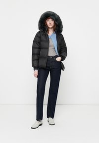 Crna puffer jakna s kapuljačom od umjetnog krzna, u paru sa sivim topom i tamnoplavim trapericama. Cipele su svijetle slip-on s crnim potplatima.