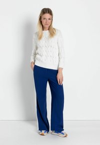 Weißer Strickpullover mit Zopf-Muster und geripptem Saum, kombiniert mit blauen weit geschnittenen Hosen mit Seitenstreifen und hellen Sneakers.