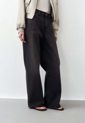 Persona che indossa jeans neri a gamba larga, giacca corta beige, top bianco e sandali marroni, in piedi davanti a uno sfondo chiaro e uniforme.