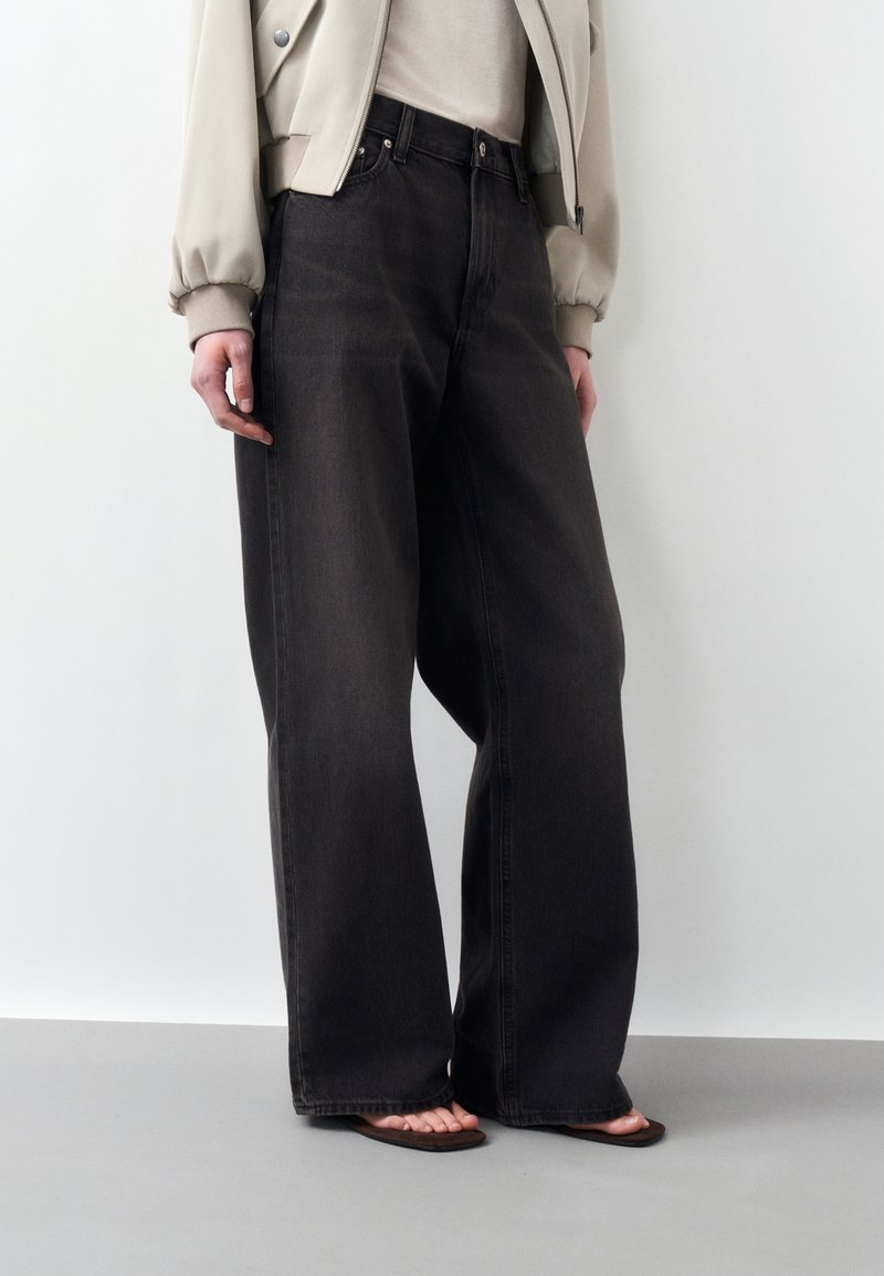 Άτομο με φαρδιά μαύρα τζιν, μπεζ cropped μπουφάν, λευκό τοπ και καφέ σανδάλια, στέκεται μπροστά σε ομοιόμορφο ανοιχτό φόντο.