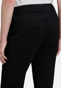 Widok z tyłu osoby noszącej czarne spodnie z elastycznym paskiem i dwiema poziomymi kieszeniami z tyłu, stojącej z zrelaksowanymi rękami.