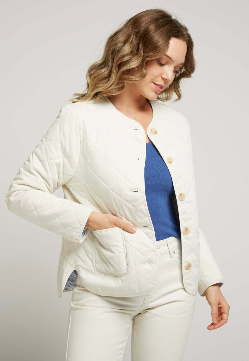 Veste crème matelassée avec col rond, fermeture à boutons et poche latérale. Portée sur un haut bleu, associée à un pantalon de couleur claire.