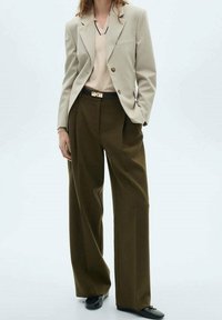 Beige Blazer mit zwei Knöpfen, kombiniert mit einem hellen Oberteil und weiten olivgrünen Hosen. Schwarze Ballerinas vervollständigen das Outfit.