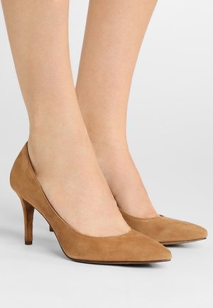Pies con zapatos de tacón alto de ante color beige con punta afilada y tacones medios delgados sobre un fondo claro.