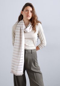 Écharpe rayée beige clair en tissu doux, drapée autour du cou, avec des bords frangés et un détail de logo. Associée à un haut neutre et un pantalon.