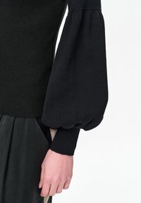 Czarny prążkowany sweter z obszernym balonowym rękawem zwężającym się przy nadgarstku, w połączeniu z czarną plisowaną spódnicą, dłoń swobodnie ułożona wzdłuż ciała.