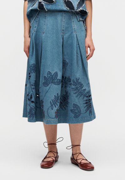 Jupe en denim plissée avec broderie florale foncée. Teinture bleu clair, longueur genou, associée à des chaussures plates marron à lacets.