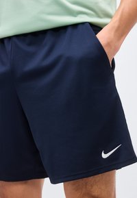Άτομο που φοράει ναυτικό-μπλε σορτς Nike με λευκό λογότυπο Swoosh, με το χέρι στην δεξιά τσέπη και ένα ανοιχτό πράσινο τοπ μερικώς ορατό.
