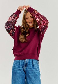 Burgundowa bluza z kwiecistymi rękawami, z okrągłym dekoltem i ściągaczami, zestawiona z niebieskimi dżinsami z wysokim stanem.