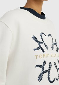 Sudadera blanca con bordado en color marino de un diseño de cuerda y texto dorado "TOMMY HILFIGER". Tiene un cuello redondo en color marino y una textura suave.